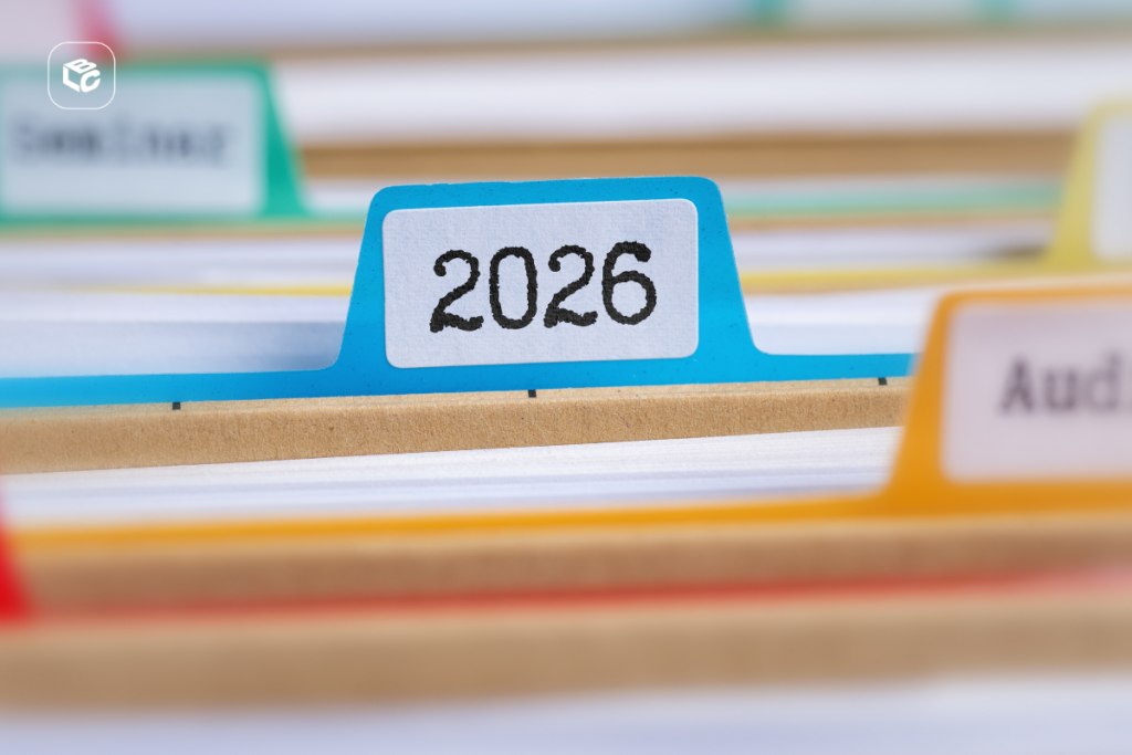 Close-up de um arquivo físico com divisórias coloridas, destacando uma aba azul identificada com o ano “2026”. Pastas e documentos organizados aparecem desfocados ao fundo, representando arquivamento, gestão documental, organização de processos e controle de registros. Logotipo da empresa visível no canto superior esquerdo.