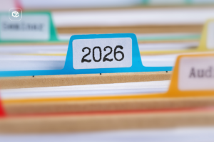 Close-up de um arquivo físico com divisórias coloridas, destacando uma aba azul identificada com o ano “2026”. Pastas e documentos organizados aparecem desfocados ao fundo, representando arquivamento, gestão documental, organização de processos e controle de registros. Logotipo da empresa visível no canto superior esquerdo.